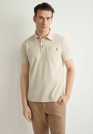 Mężczyzna ubrany w beżową koszulkę polo z kieszenią na piersi i brązowe spodnie, stojący z ręką w kieszeni na białym tle.