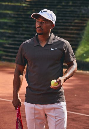 Mannelijke tennisser in een witte pet en korte broek die een bal en racket vasthoudt, staande op een gravelbaan met wazige stappen op de achtergrond.