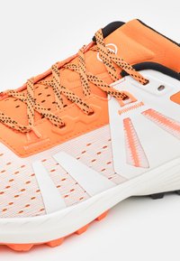 Pomarańczowy i biały but sportowy z siateczkową cholewką, perforowanym wzorem i teksturowanymi akcentami. Zawiera kontrastujące czarne elementy i mocne sznurówki.