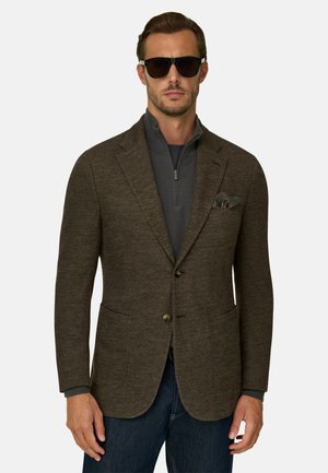 Homme portant des lunettes de soleil foncées, un blazer marron texturé avec un mouchoir de poche, un pull gris foncé à col zippé et un jean bleu foncé, debout sur un fond blanc.