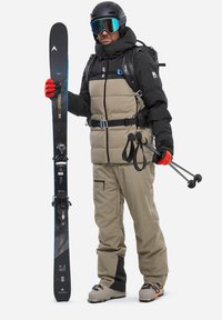 Skiausrüstung umfasst eine schwarze und beige isolierte Jacke, beigefarbene Skihosen, rote Handschuhe, Skibrille, schwarze Skier und Trekkingstöcke.