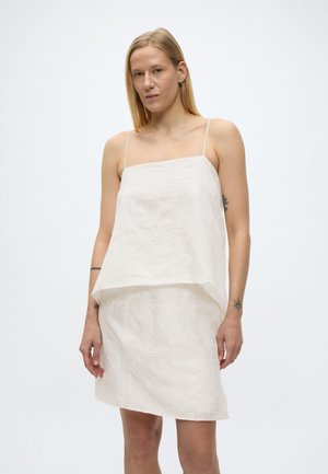 Blonde woman with straight hair wears a sleeveless white dress with thin straps and subtle embroidered patterns, standing against a plain background.