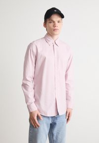 Lichtroze overhemd met lange mouwen, voorzien van een klassieke kraag en een relaxte pasvorm, gecombineerd met blauwe denimjeans en een zwarte pet.