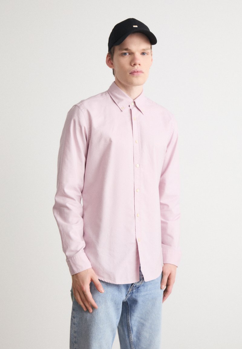 Lichtroze overhemd met lange mouwen, voorzien van een klassieke kraag en een relaxte pasvorm, gecombineerd met blauwe denimjeans en een zwarte pet.