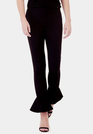 Femme portant un pantalon noir ajusté avec des ourlets évasés à volants et des sandales noires à talons hauts ouvertes, debout devant un fond blanc.