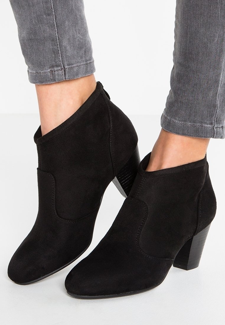 Bottines noires en daim à talons carrés portées avec un jean gris retroussé sur pieds nus.