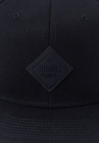 Melna cepure ar strukturētu audumu, plakanu maliņu un taisnstūrveida reljefu logotipa uzlīmi ar zīmolu "DJINNS Supply Co." centrā.