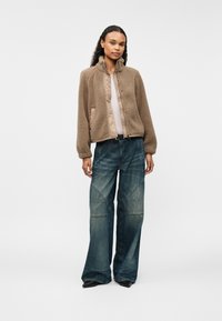 Chaqueta de felpa marrón con cremallera y un cuello alto, combinada con jeans de pierna ancha azul con un patrón cosido. Diseño texturizado y casual.