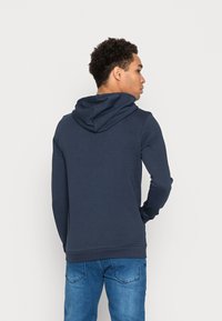 Felpa blu navy con cappuccio regolabile, realizzata in materiale morbido. Presenta polsini e orlo a coste, con un design elegante e attillato. I jeans sono di un blu chiaro.