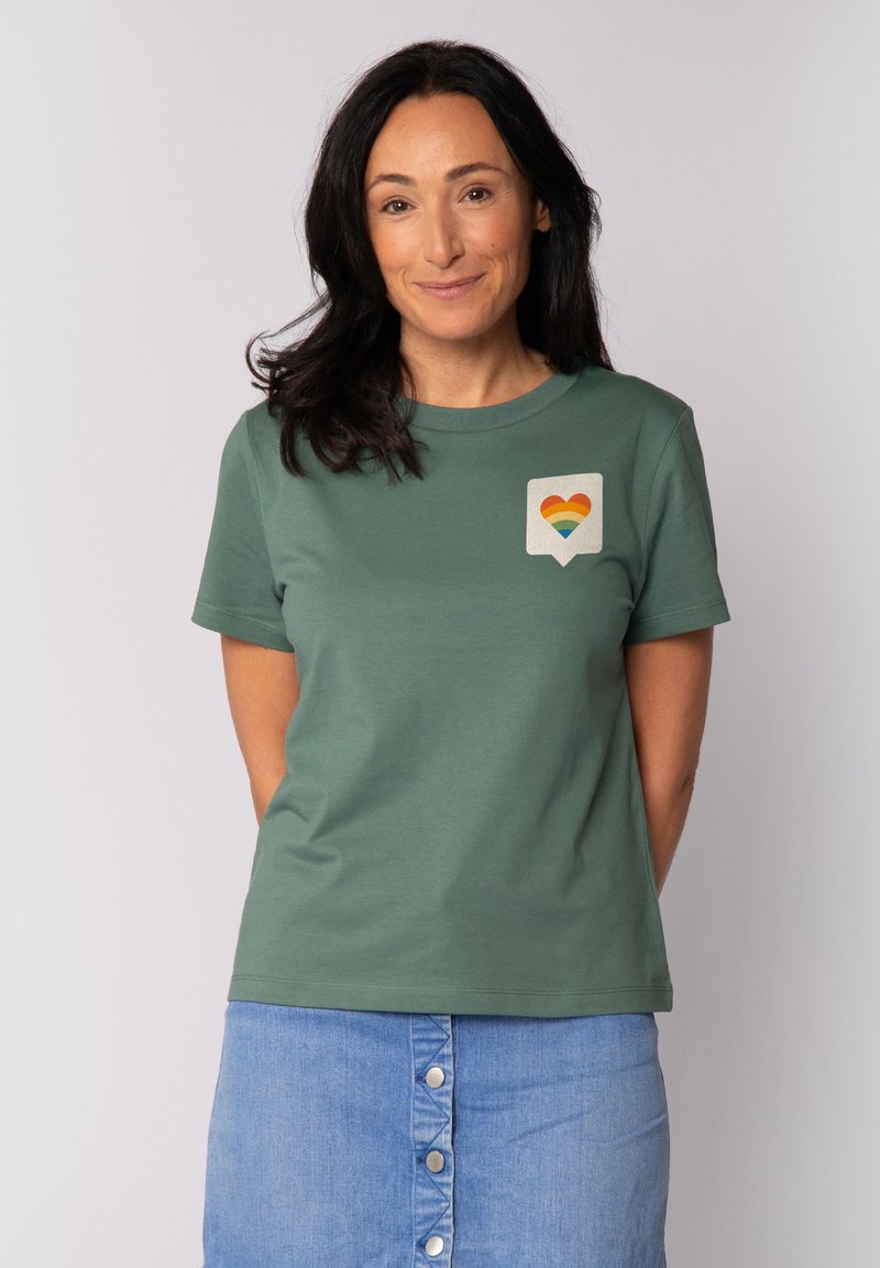 T-shirt en coton vert avec un patch représentant un cœur arc-en-ciel sur la poitrine gauche. Le t-shirt a des manches courtes et un col rond.