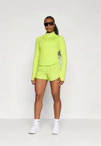 Sportinis aprangos komplektas ryškios žalios spalvos; ilgomis rankovėmis marškinėliai su pusiau užtrauktuku, derinami su atitinkančiomis šortais. Audinys atrodo lygus, sportiško stiliaus ir prigludusio silueto.