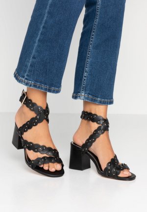 Füße, die schwarze geflochtene Riemchen-High-Heel-Sandalen tragen, kombiniert mit blauen gekürzten Jeans, stehend auf einer weißen Oberfläche.