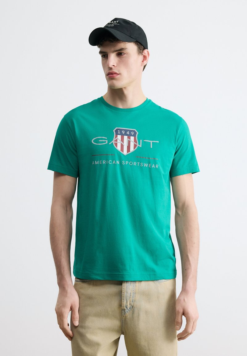 Νεαρός άνδρας που φοράει πράσινο t-shirt GANT με λογότυπο ασπίδας και κείμενο, μπεζ παντελόνι και μαύρο καπέλο, στέκεται μπροστά σε απλό φόντο.