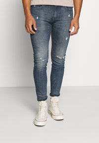 Hombre que lleva unos vaqueros ajustados desgastados y rasgados de color azul y zapatillas deportivas de caña alta blancas, de pie contra un fondo claro y sencillo.
