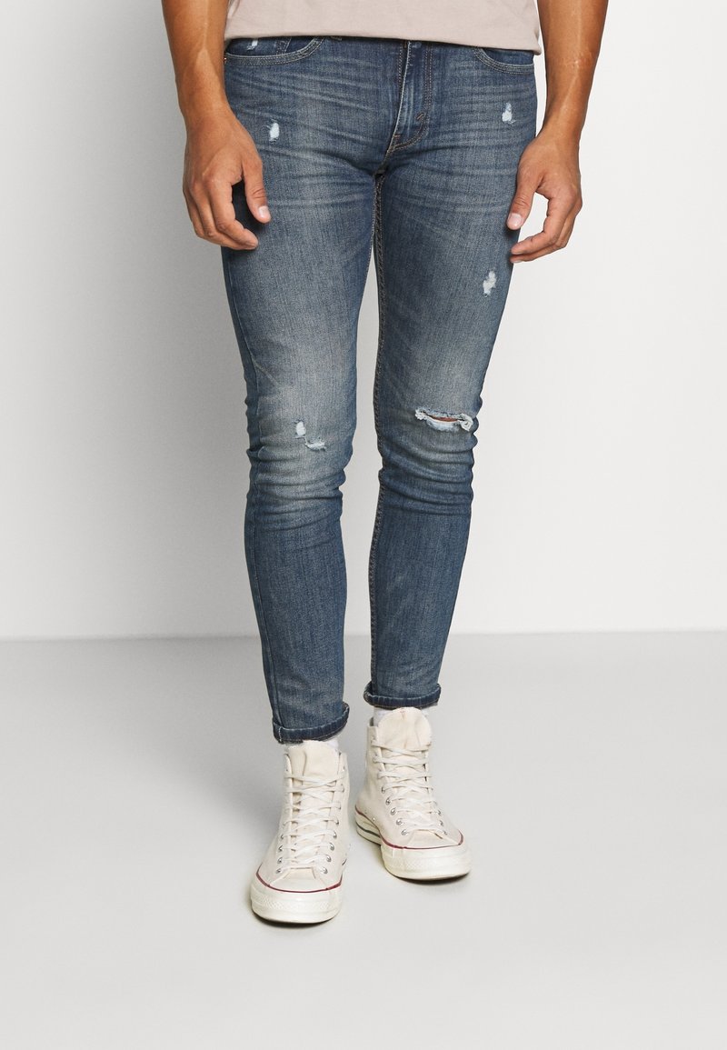 Hombre que lleva unos vaqueros ajustados desgastados y rasgados de color azul y zapatillas deportivas de caña alta blancas, de pie contra un fondo claro y sencillo.