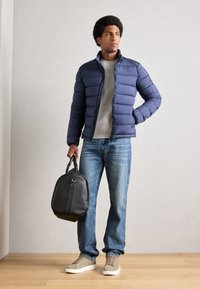 Veste matelassée marine, chemise grise, jeans bleus et baskets beiges. Sac noir avec poignées et détails de couture tenu à la main. Design simple et moderne.