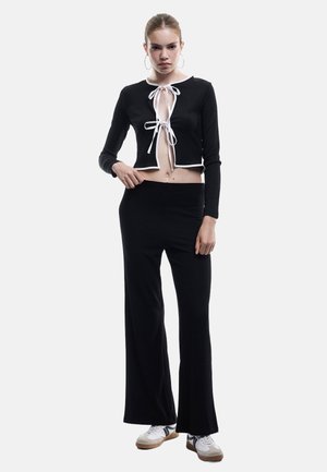 Schwarzes geripptes Croptop mit weißem Saum und Bänder-Tie-Verschluss, kombiniert mit schwarzen weiten Hosen. Das Model trägt weiße und graue Sneakers.