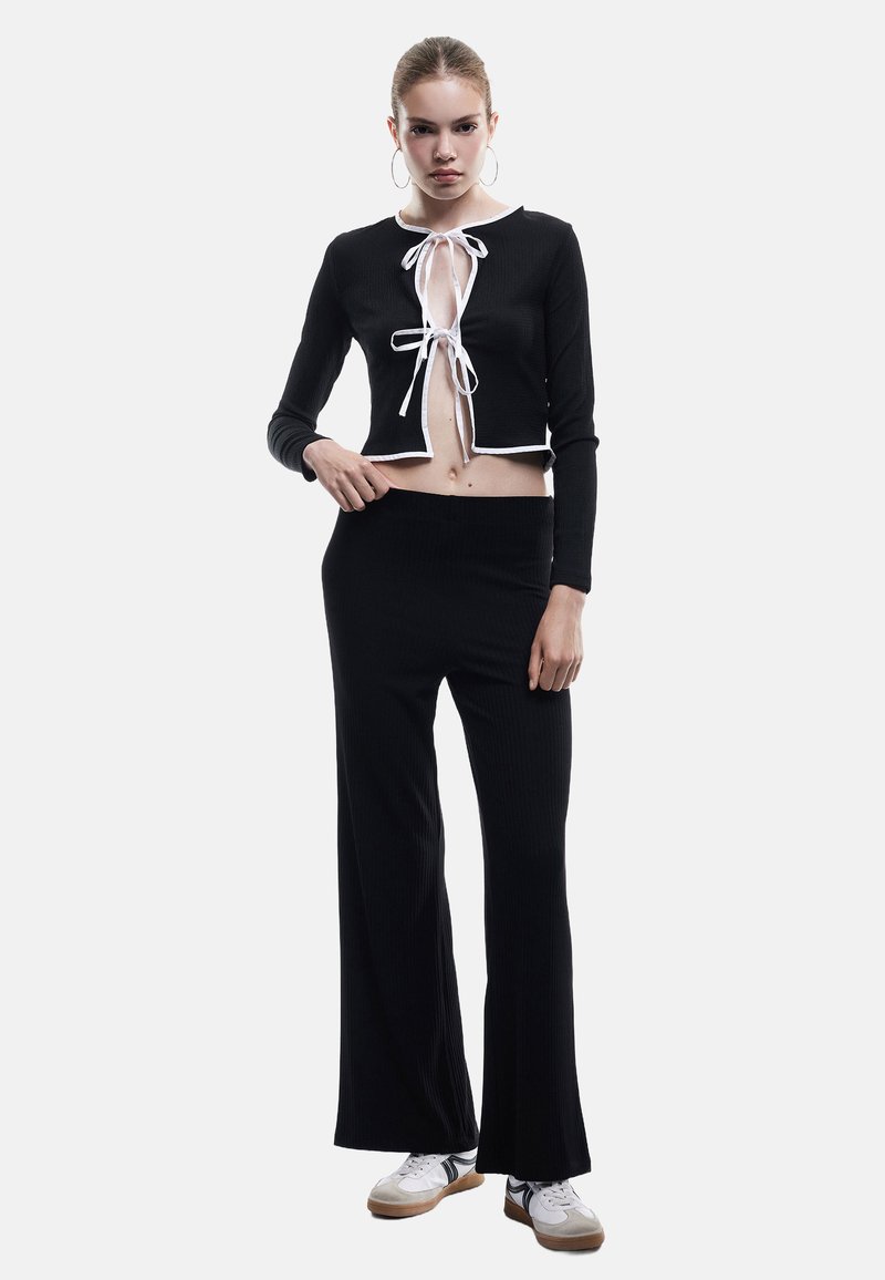 Svart ribbat crop top med vit kant och snörning, i kombination med svarta vida byxor. Modell bär vita och grå sneakers.