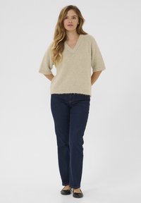 Pull en tricot beige à col en V et manches courtes, associé à un jean droit bleu foncé et des chaussures plates noires.