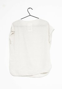 Blusa leggera di colore crema con un sottile motivo geometrico, scollo rotondo e maniche corte, esposta su un appendiabiti nero.