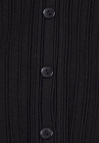 Svart ribbad trikå med sex runda svarta knappar längs framsidan. Texturen är upphöjd med vertikala linjer, vilket ger visuellt intresse.