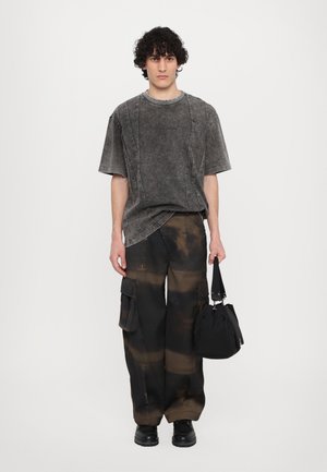 Gray oversized t-shirt with visible stitching, paired with loose black cargo pants featuring tan paint splatters and multiple pockets, holding a black bag.