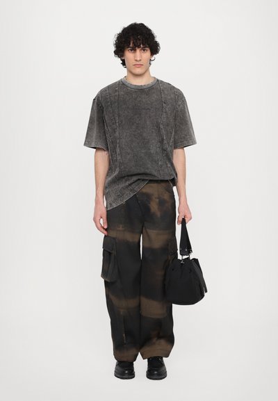 T-shirt gris surdimensionné avec des coutures visibles, associé à un pantalon cargo noir ample présentant des éclaboussures de peinture beige et de multiples poches, tenant un sac noir.