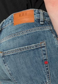Jeans de mezclilla azul con una etiqueta de cuero beige que dice "R.D.D" en la cinturilla. Se pueden ver costuras rojas notables y bolsillos traseros estándar.