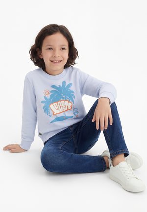 Bambino con capelli ricci seduto a gambe incrociate che indossa una felpa celeste chiaro con stampa di palme, jeans blu e scarpe da ginnastica bianche su sfondo bianco.