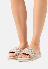 Pletené sandály ve světle béžové barvě, vyrobené z měkkého materiálu. Mají silné tkané pruhy a plochou, polstrovanou podrážku s jemným švem.