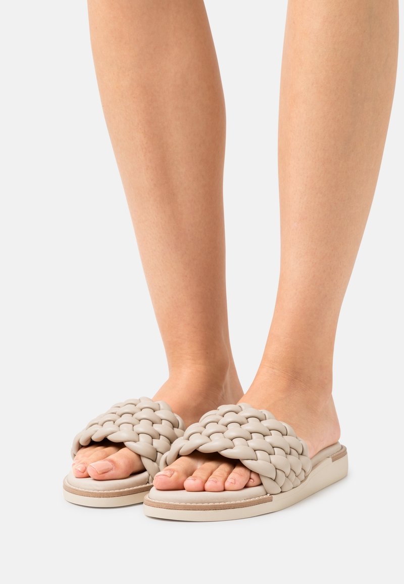 Pletené sandály ve světle béžové barvě, vyrobené z měkkého materiálu. Mají silné tkané pruhy a plochou, polstrovanou podrážku s jemným švem.