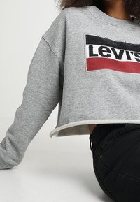 Felpa grigia corta con logo Levi's testurizzato in nero, rosso e bianco. Presenta un colletto e un orlo a coste, abbinata a jeans neri.