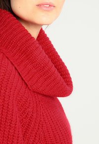 Rode gebreide trui met een brede, omgevouwen col. Gelaagd patroon met een geribbelde structuur, gemaakt van zacht, dik garen.