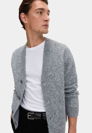 Lys grå, tekstureret cardigan med V-hals, knaplukning og ribbede manchetter. Båret over en hvid skjorte og parret med mørke bukser.