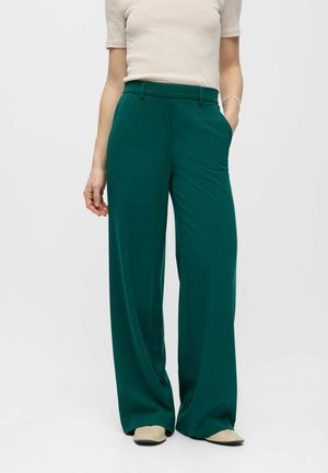 Personne portant un pantalon large taille haute couleur sarcelle avec poches latérales et chaussures pointues beige, assorti à un haut beige clair à manches courtes.