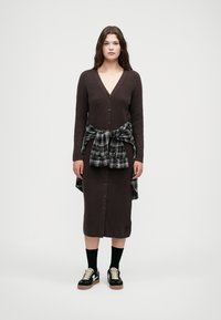 Καφέ ριγωτό πλεκτό φόρεμα με V-λαιμόκοψη και κουμπιά κατά μήκος του μπροστινού μέρους, συνδυασμένο με μία μαύρη και λευκή καρό πουκαμίσα δεμένη γύρω από τη μέση. Μαύρα αθλητικά παπούτσια με κρεμ λεπτομέρειες.