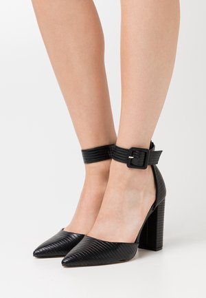 Chaussures noires à talons hauts avec un bout pointu, un matériau texturé et une sangle de cheville avec une boucle carrée ; le talon bloc ajoute de la stabilité.