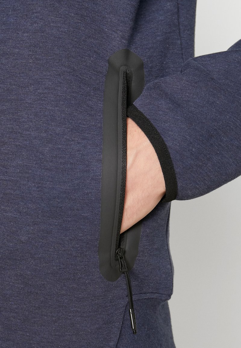 Sweatshirt marine en tissu texturé, comportant une poche zippée avec une bordure noire. La poche est partiellement ouverte, révélant une main.