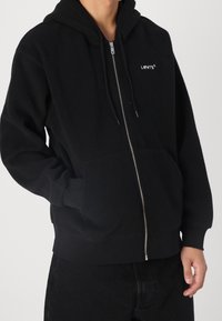 Svart fleecehoodie med dragkedja och framfickor. Har en liten vit logotyp på vänster sida samt ribbade muddar och fåll.