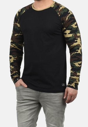 Homme portant une chemise noire à manches longues avec des manches camouflages et un jean gris clair, debout avec une main dans la poche.