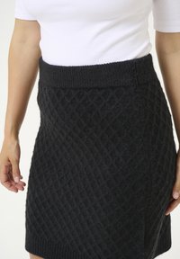 Gonna nera lavorata a maglia con un motivo a diamante, vita a costine e una texture morbida. L'orlo presenta una leggera inclinazione per un look contemporaneo.