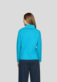 Frau mit hellbraunem Haar, die ein leuchtend blaues Langarmoberteil und eine marineblaue Hosen mit Nadelstreifen trägt, mit dem Rücken zu einem einfarbigen Hintergrund gewandt.