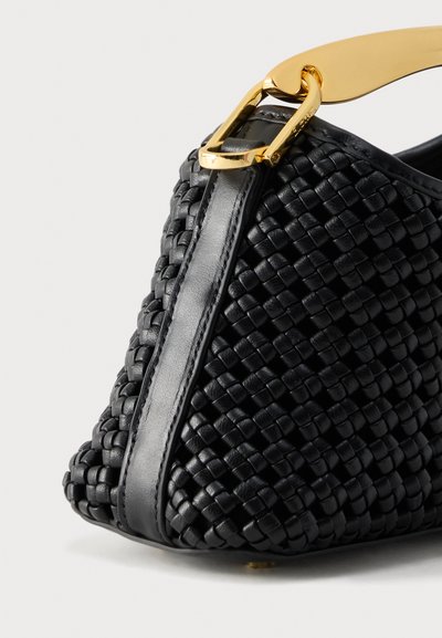 Μαύρη υφασμάτινη γωνία δερμάτινης τσάντας με λωρίδα από λείο δέρμα και χρυσό μεταλλικό κούμπωμα και λεπτομέρεια λουριού σε ουδέτερο φόντο.