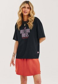 Čierne oversized tričko s potlačou "HOLD YOUR HORSES" v ružovej farbe, v kombinácii s voľnou oranžovou sukňou. Výrazné ploché švy a krátke rukávy.