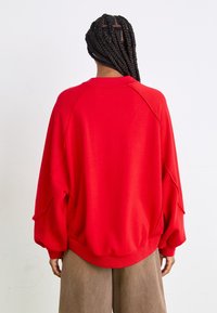 Red oversized sweatshirt with ribbed hem and cuffs, featuring a relaxed fit and raglan sleeves, paired with brown wide-leg pants.