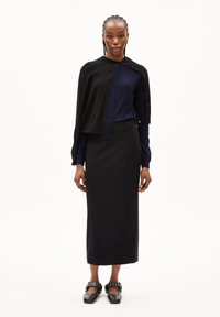 Zwarte cardigan over een blauwe langemouwen top, gecombineerd met een zwarte rok. Zwarte schoenen met zilveren hardware accentueringen maken de outfit compleet.