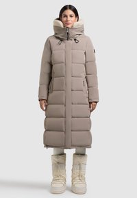 Palton pufos lung bej cu design matlasat, guler înalt și fermoar dublu. Glugă tapitată cu fleece și buzunare laterale. Asortat cu cizme albe de zăpadă.