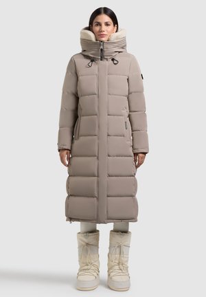 Dolga bež puhovka s prešanim vzorcem, visokim ovratnikom in dvojnim zadrgo. Kapuca obložena s flisom in stranska žepa. V kombinaciji z belimi snežnimi čevlji.