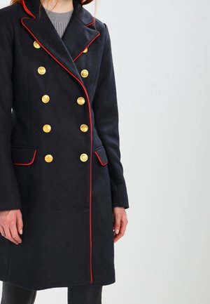 Kvinde iført en marineblå dobbeltradet frakke med guldknapper og røde pipingdetaljer, stående mod en ensfarvet baggrund.