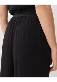 Czarne spodnie mają gładką fakturę, szeroką elastyczną talię oraz dwie tylne kieszenie. Ich fason jest luźny i zwiewny.